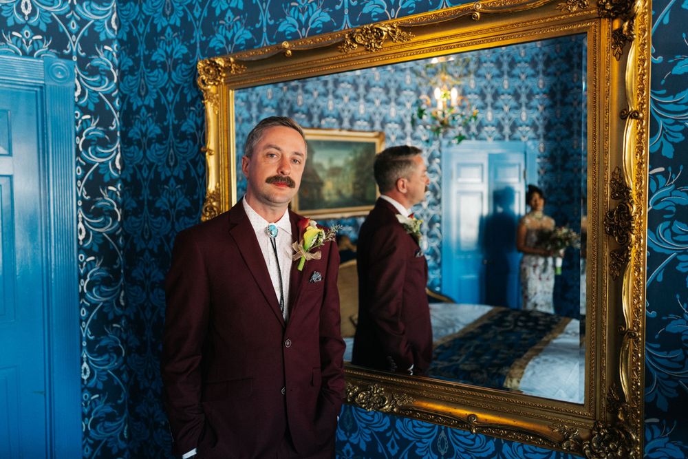 Person in burgundy suit and boutonniere poses beside ornate gold mirror against blue damask wallpaper.