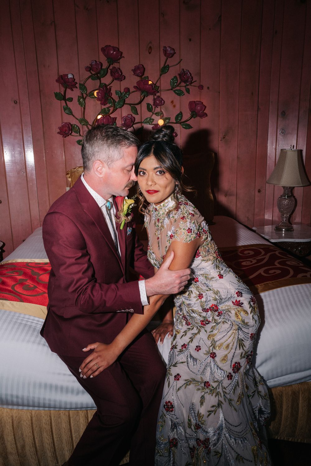 A couple in formal attire pose together on a bed, with her wearing a floral embroidered gown and him in a burgundy suit.