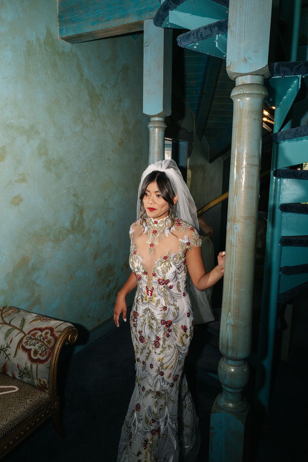 A person wearing an elegant white floral embroidered mermaid wedding dress poses in a vintage-style interior setting.