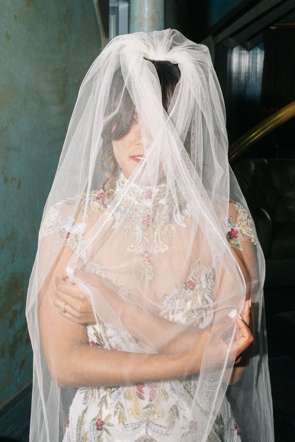 A bride wearing a delicate sheer veil and embroidered white dress stands in soft lighting.