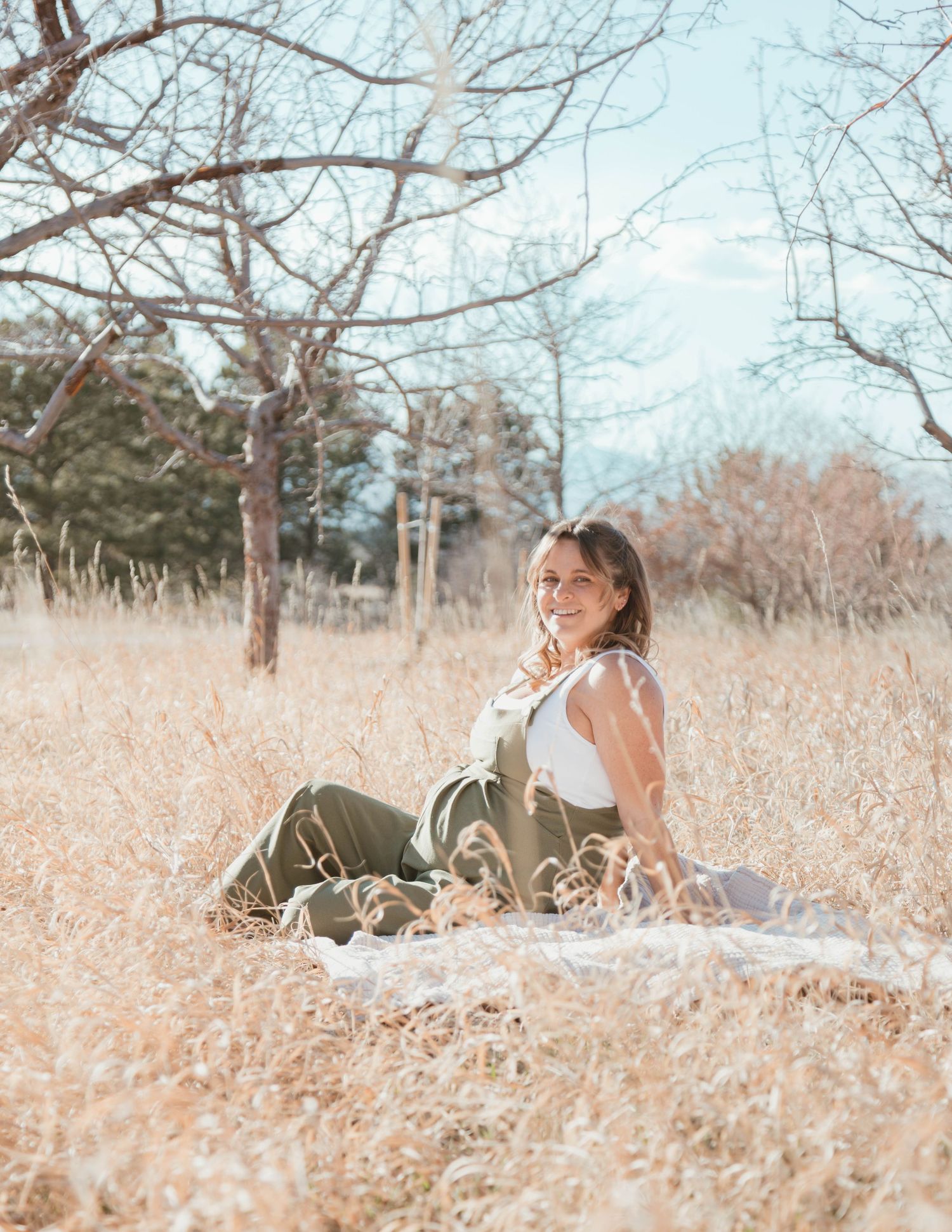 Dreamy outdoor maternity photo shoot in a sunlit field with dried grass and bare winter trees.