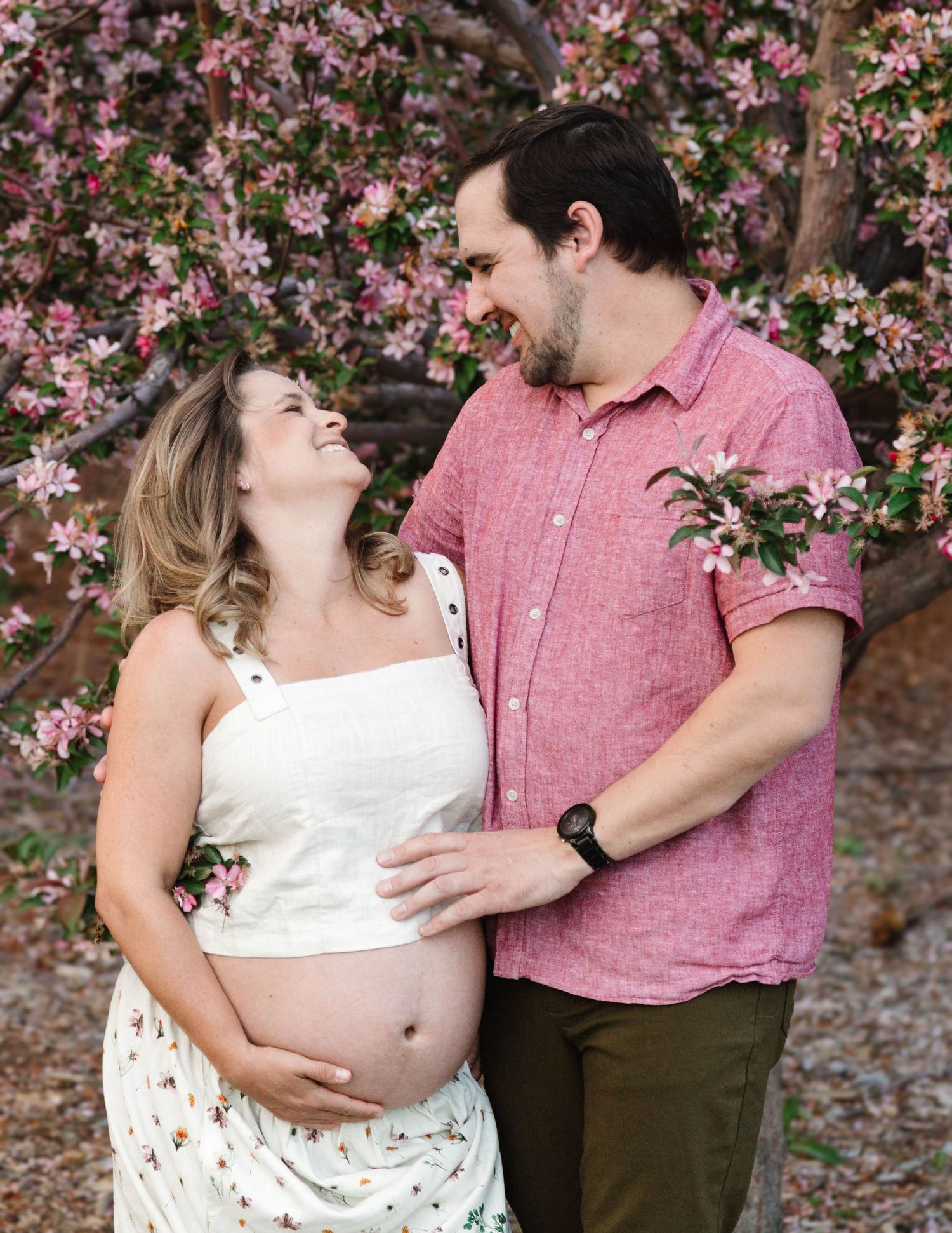 Couple in pink and white outfits share tender moments in front of blooming pink flowers during maternity photoshoot.