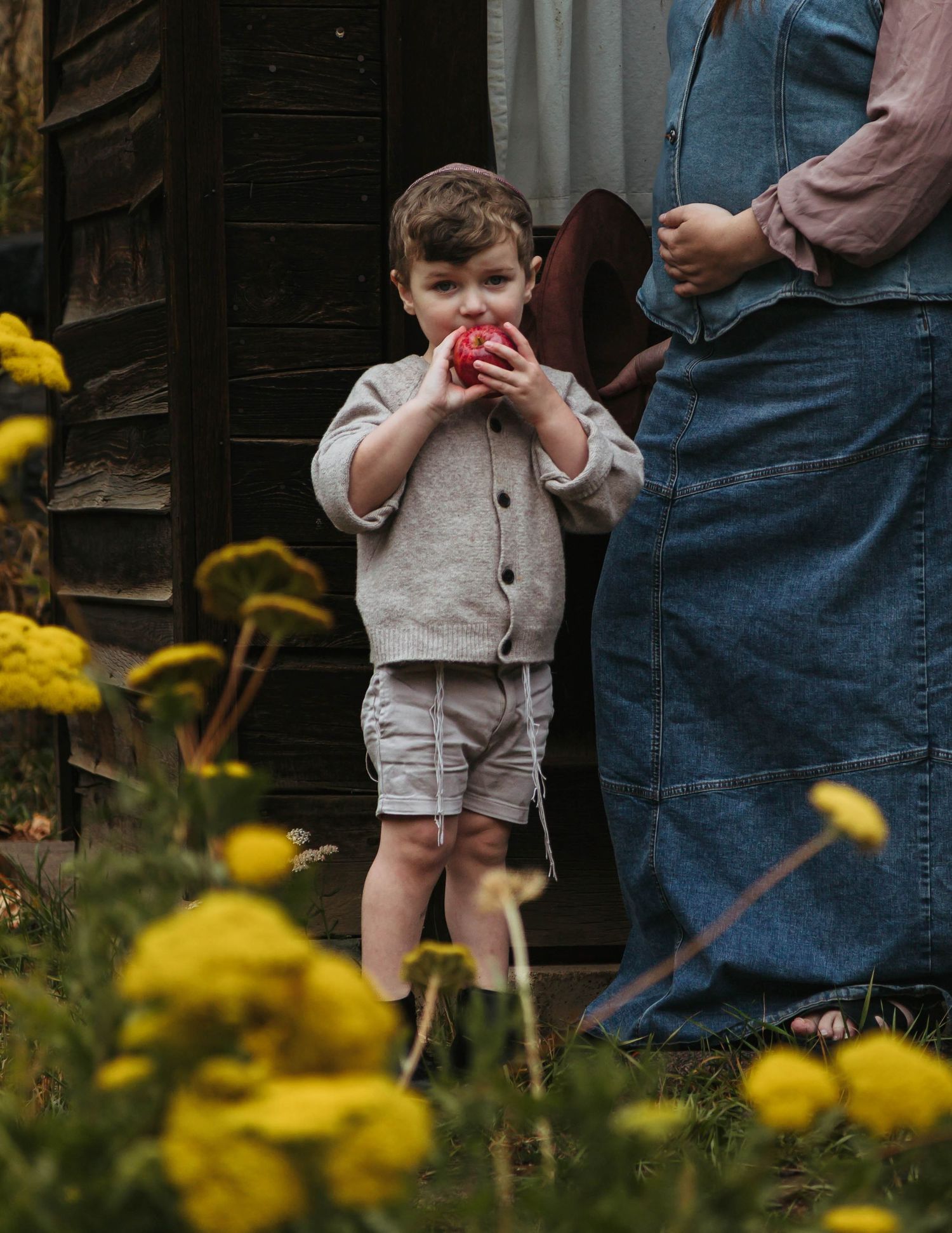 A child in beige shorts stands in a garden with yellow flowers while someone in denim jeans stands nearby.