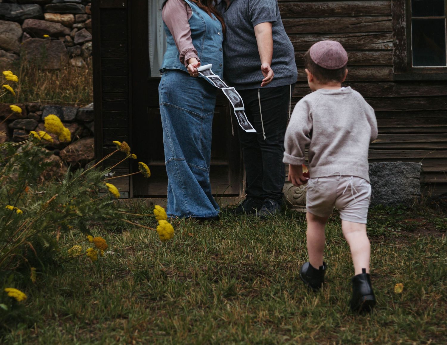 Two people walk together past a rustic wooden cabin with yellow wildflowers blooming in the foreground.