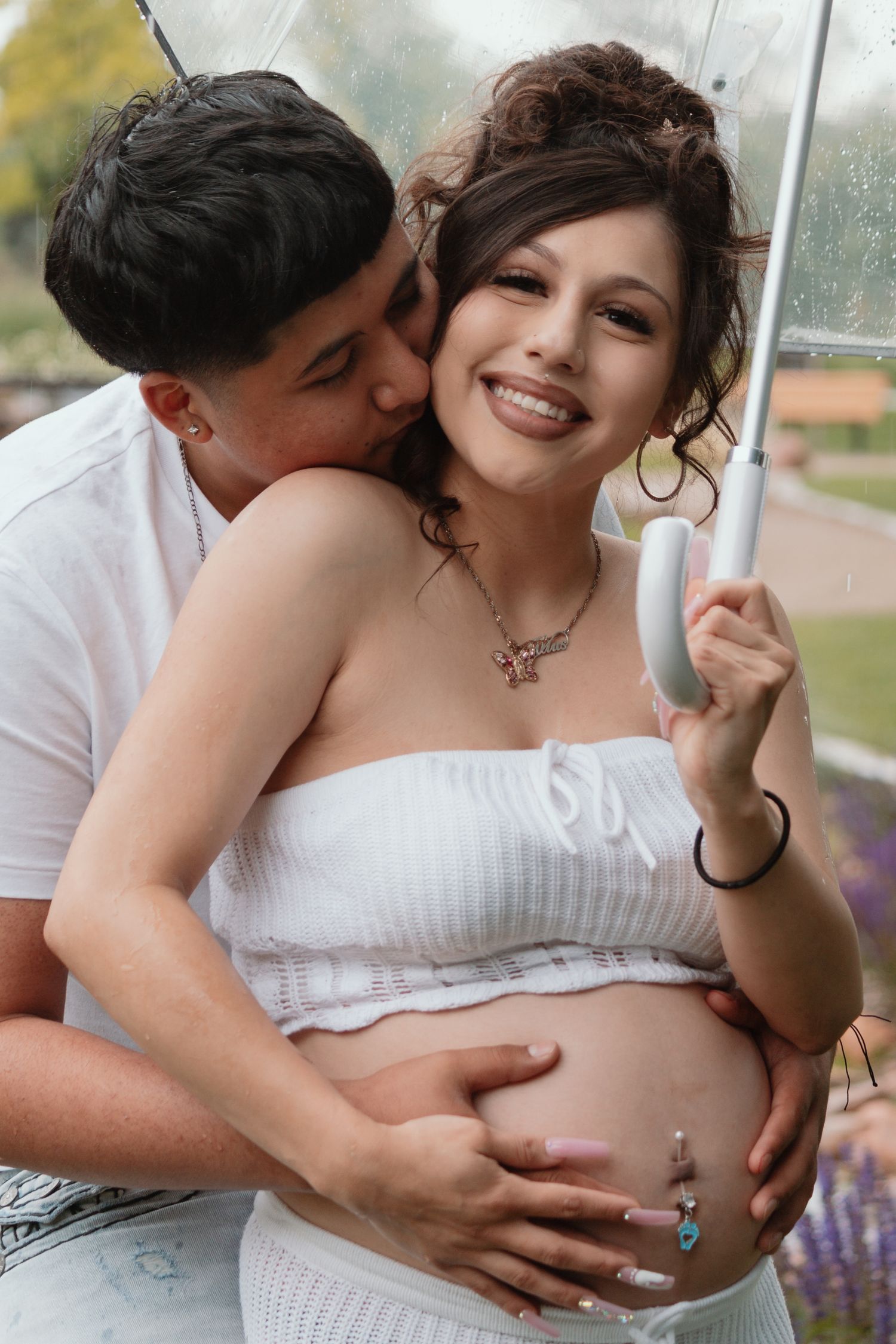 A tender maternity photo outdoors with a couple embracing under an umbrella in white attire.
