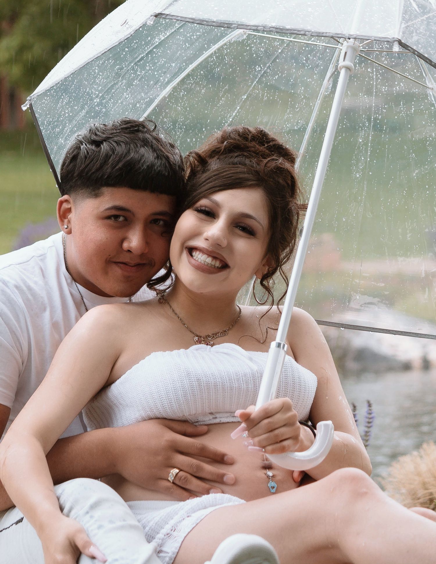 A couple shares an intimate moment under a clear umbrella in the rain, dressed in wedding attire.