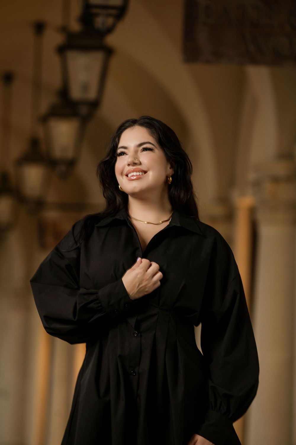 A person in a black dress with long sleeves smiles while standing in front of an archway with vintage lanterns.