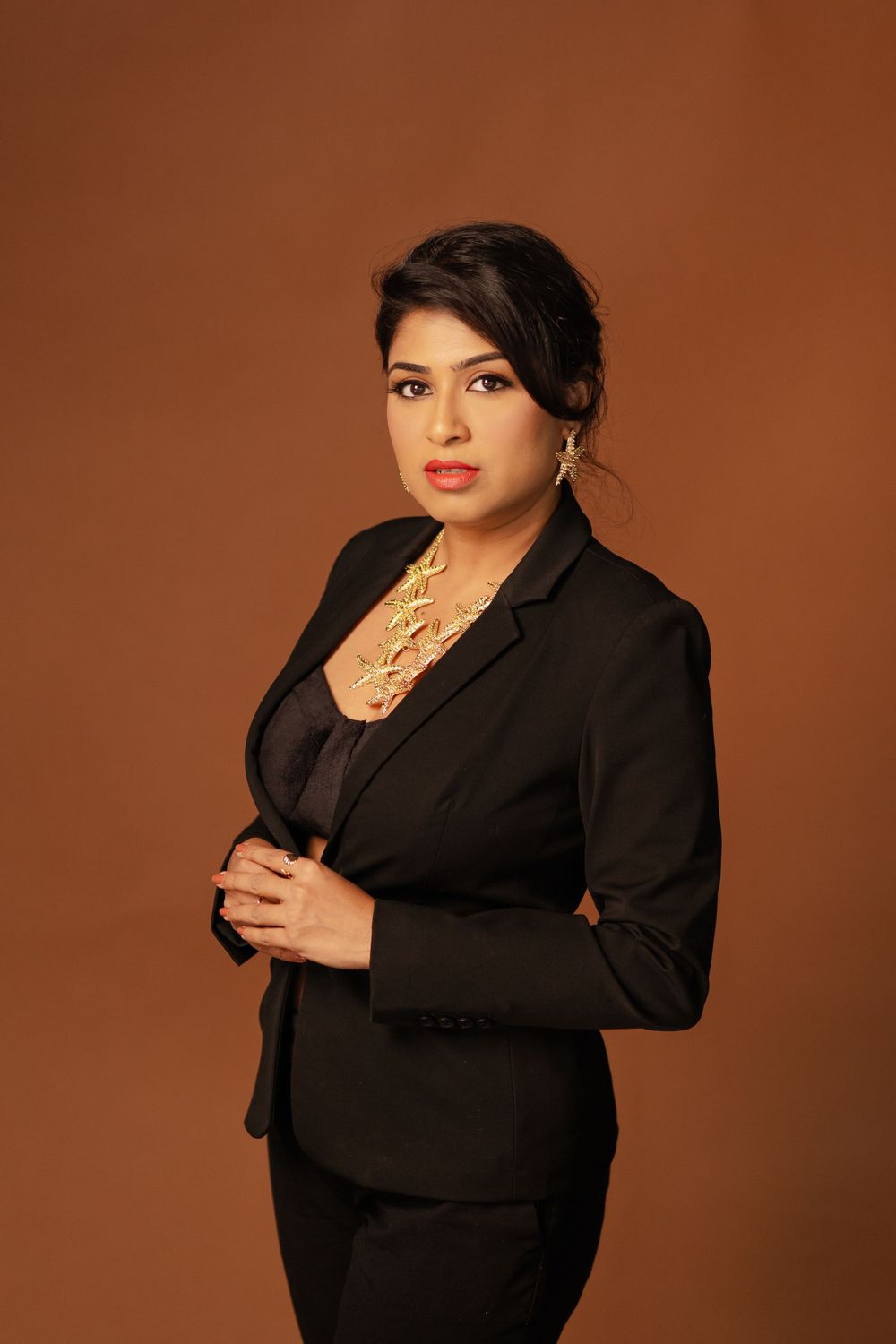 Professional portrait against brown backdrop showing person in black business suit with statement necklace.