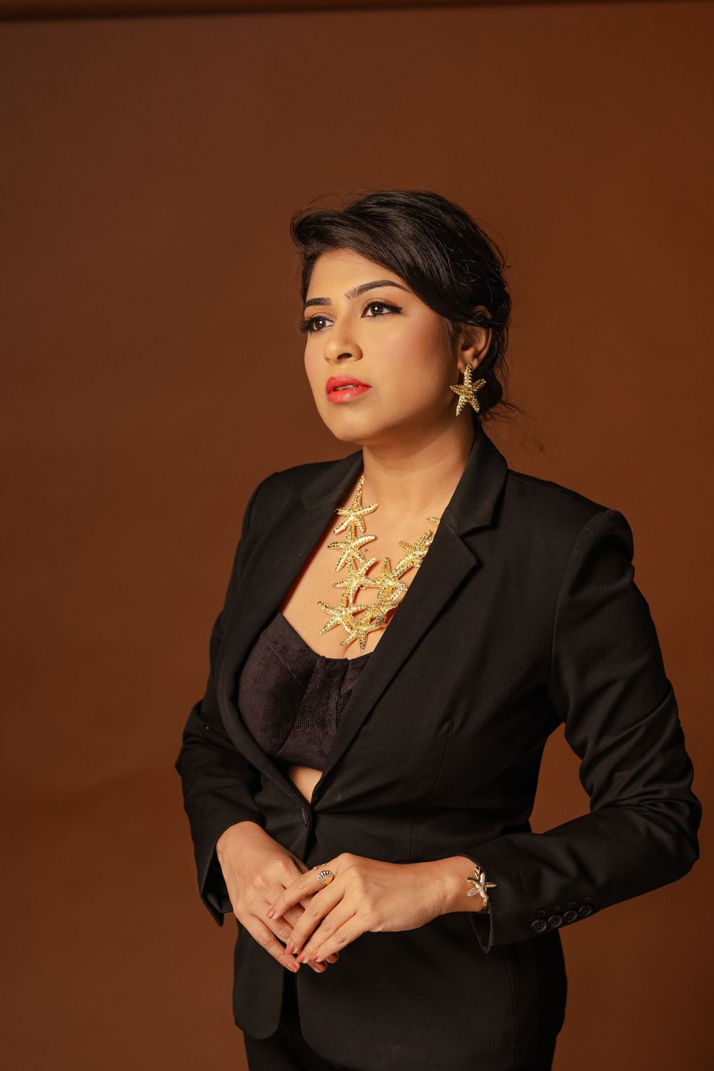 Professional portrait against brown background showing person in black blazer with statement gold necklace and earrings.