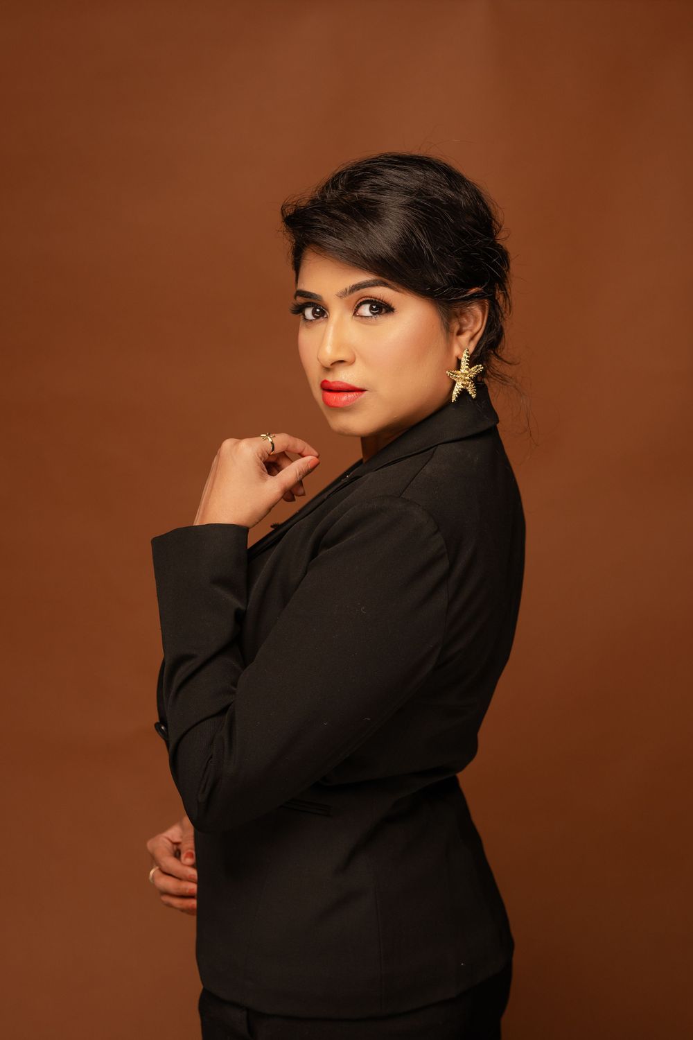 Professional studio portrait of someone in a black blazer against a brown backdrop with dramatic lighting and gold earrings.