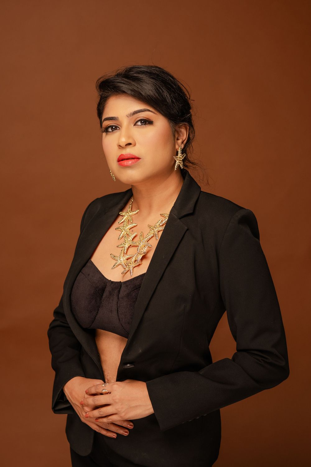 Professional studio portrait with black blazer and statement necklace against warm brown background.