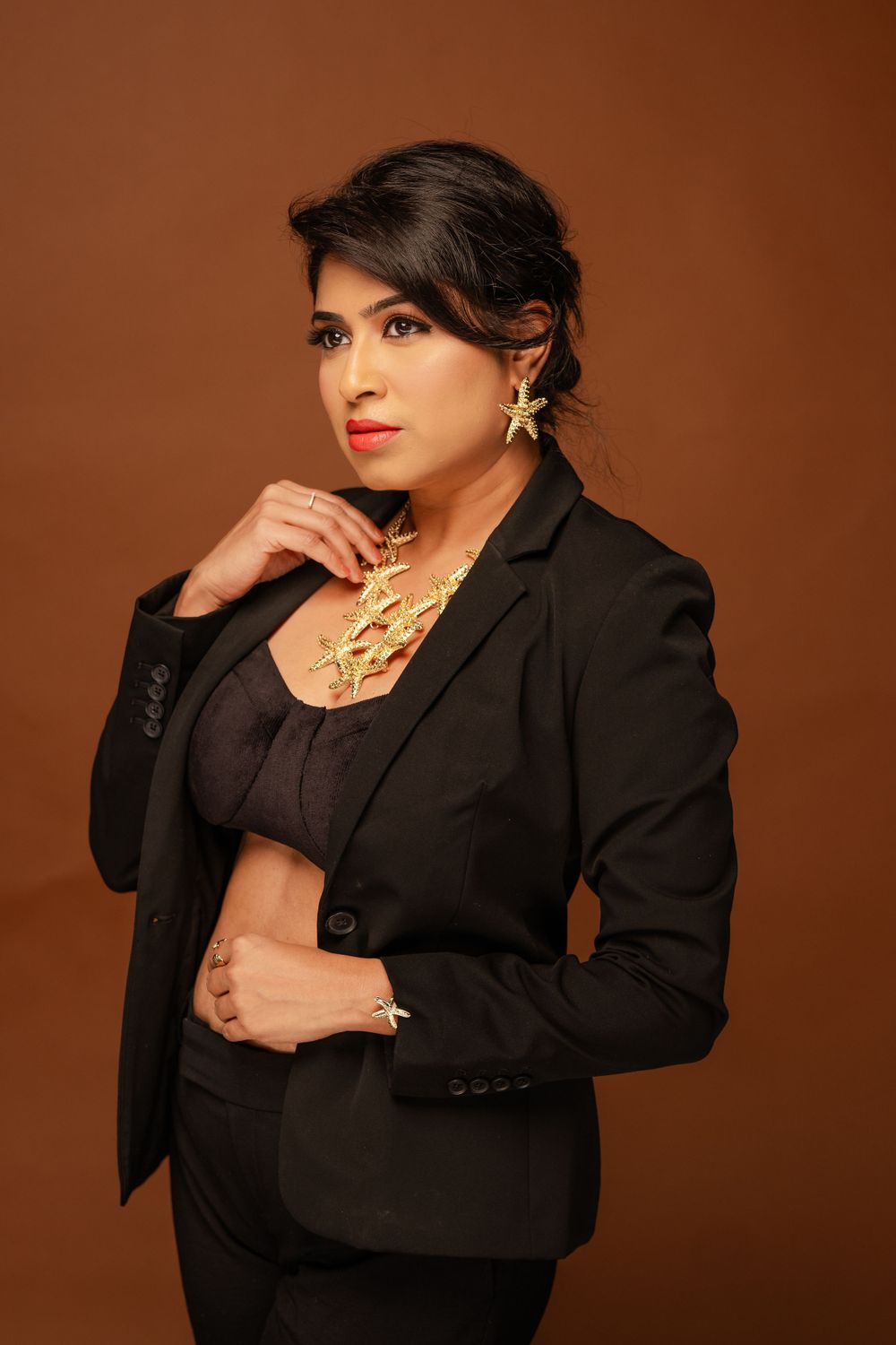Professional portrait against brown background showing elegant black business suit with gold statement necklace and earrings.