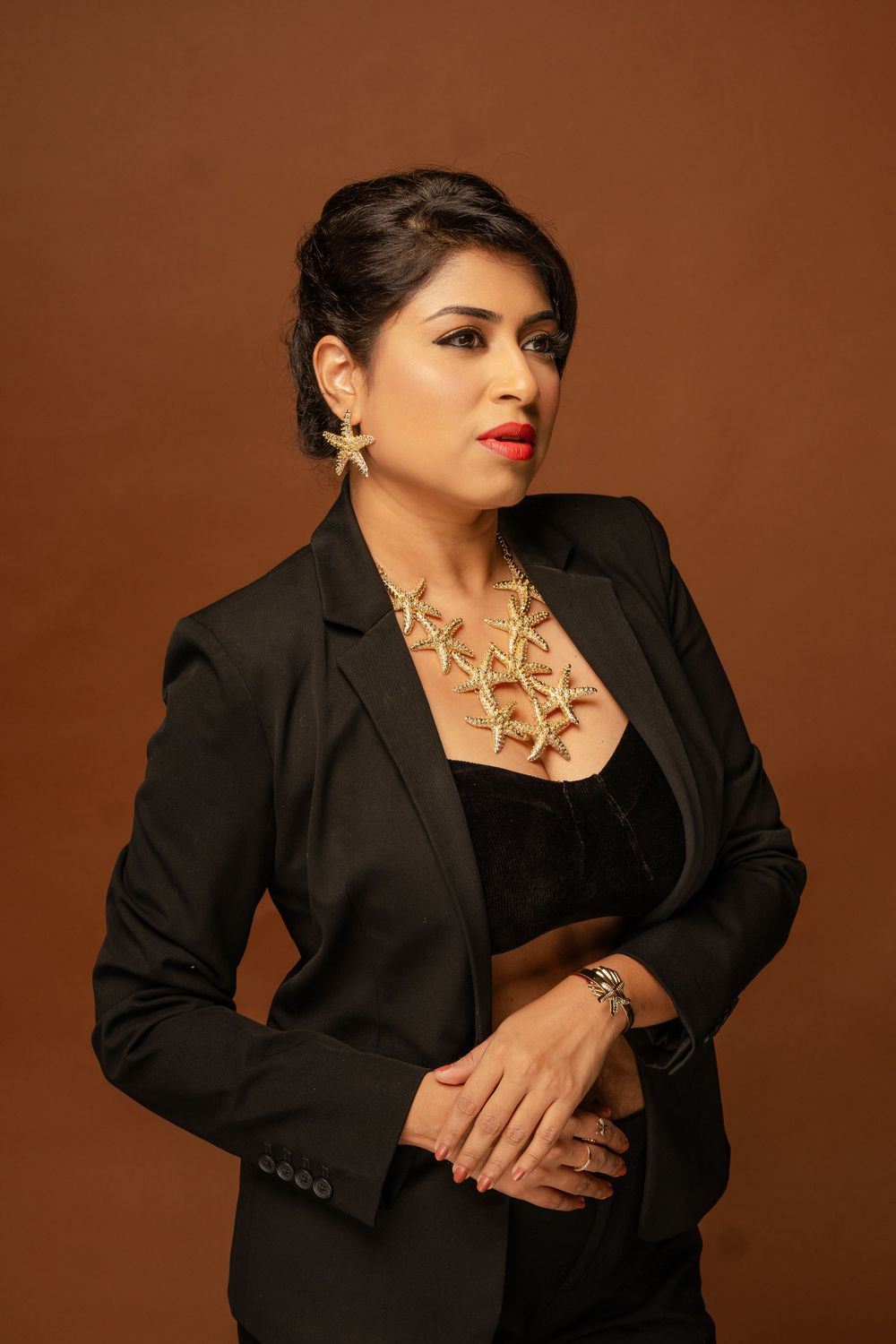 Professional portrait of a person in a black blazer and statement necklace against a brown backdrop.
