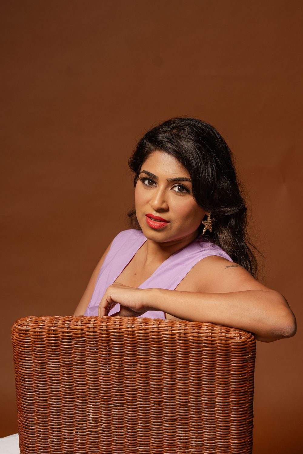 Woman in lavender top poses elegantly against woven wicker chair on brown background.