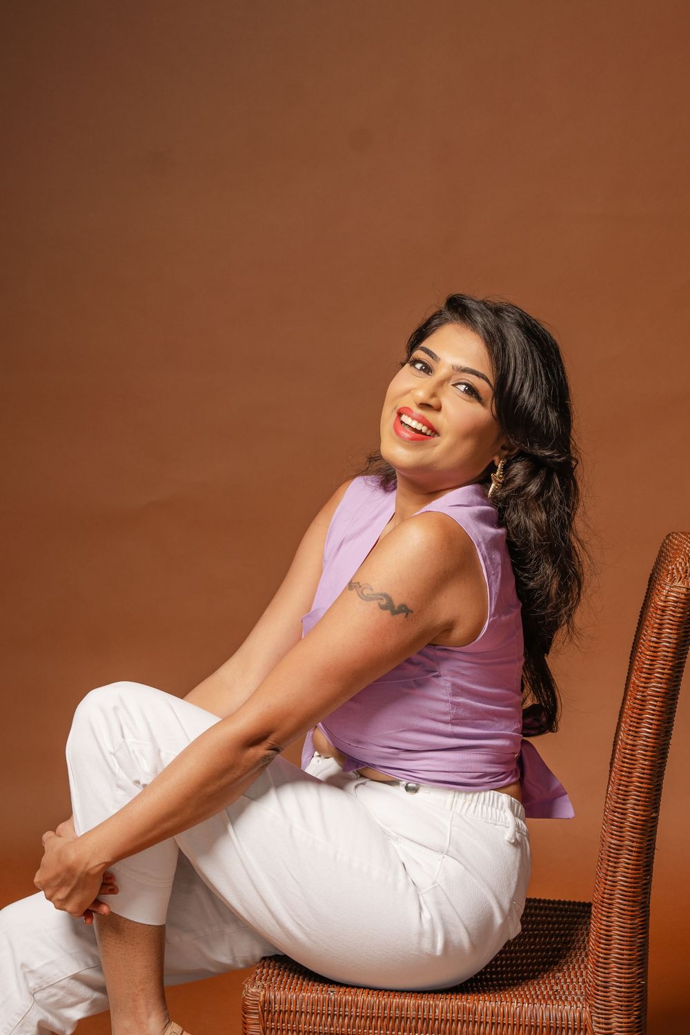 A person in a lavender tank top and white pants poses cheerfully on a wooden chair against a brown backdrop.