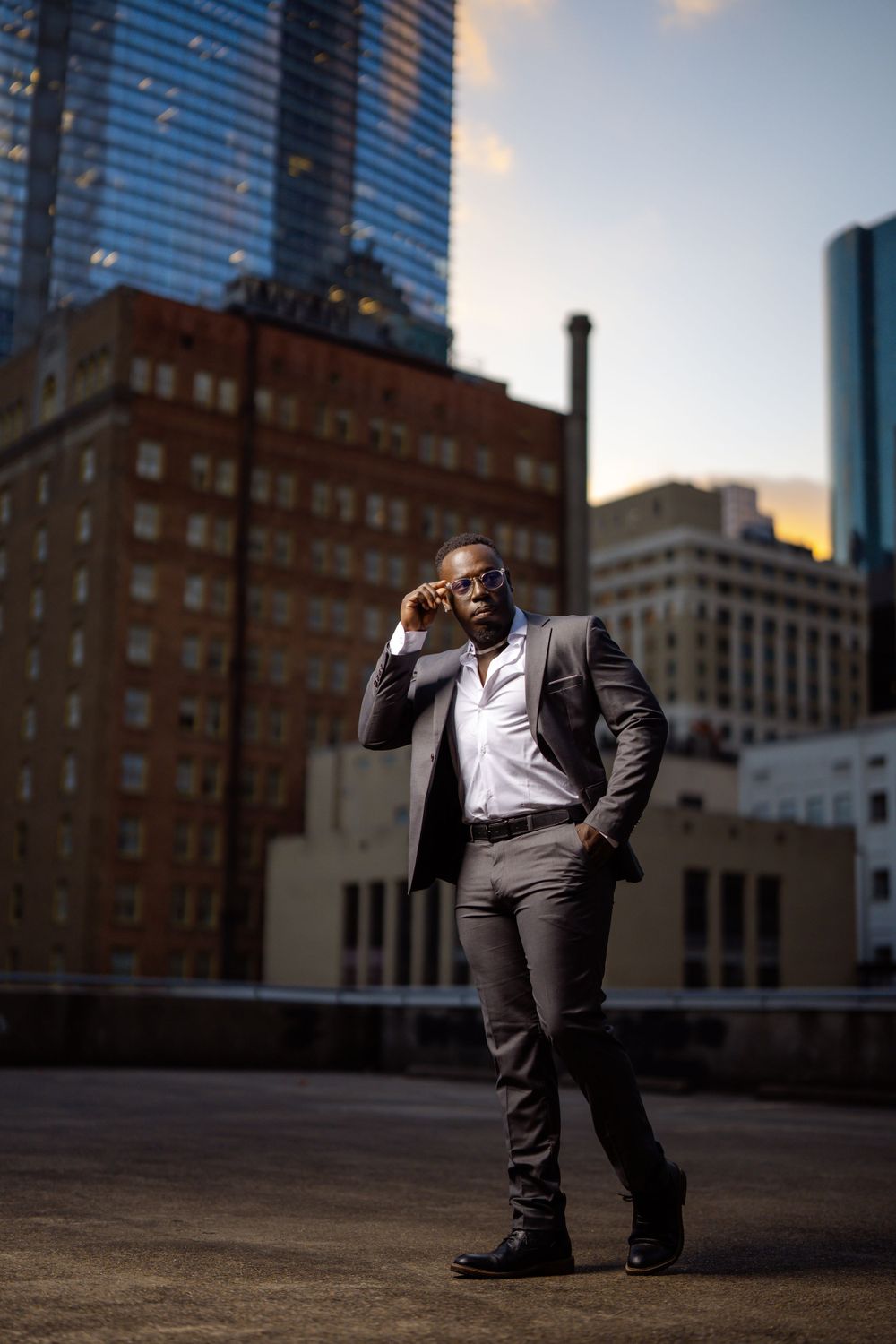 A well-dressed person in a gray suit poses confidently on a rooftop with urban skyscrapers in the background at sunset.