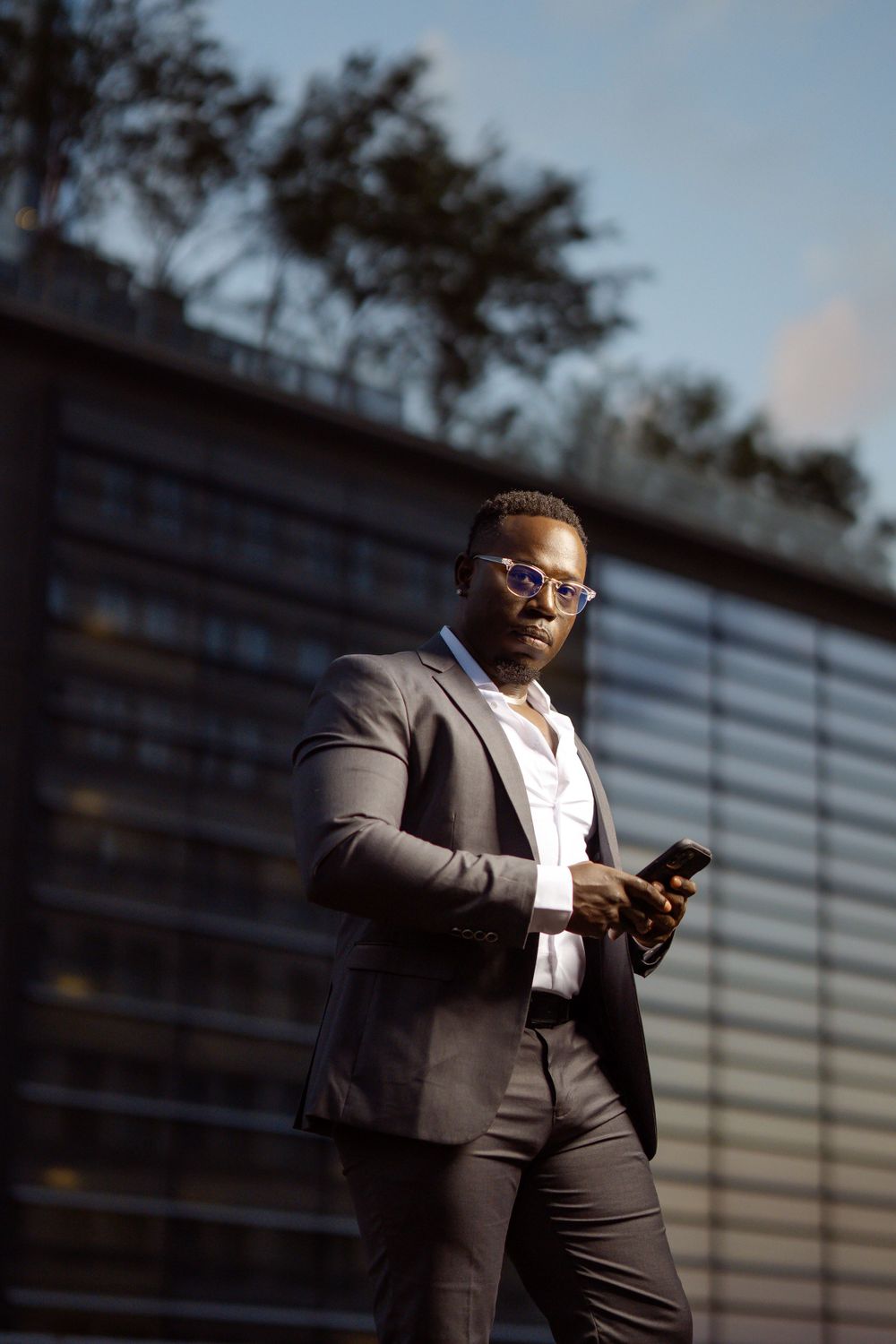 Professional in a gray suit and glasses checks smartphone while standing outside modern glass building.