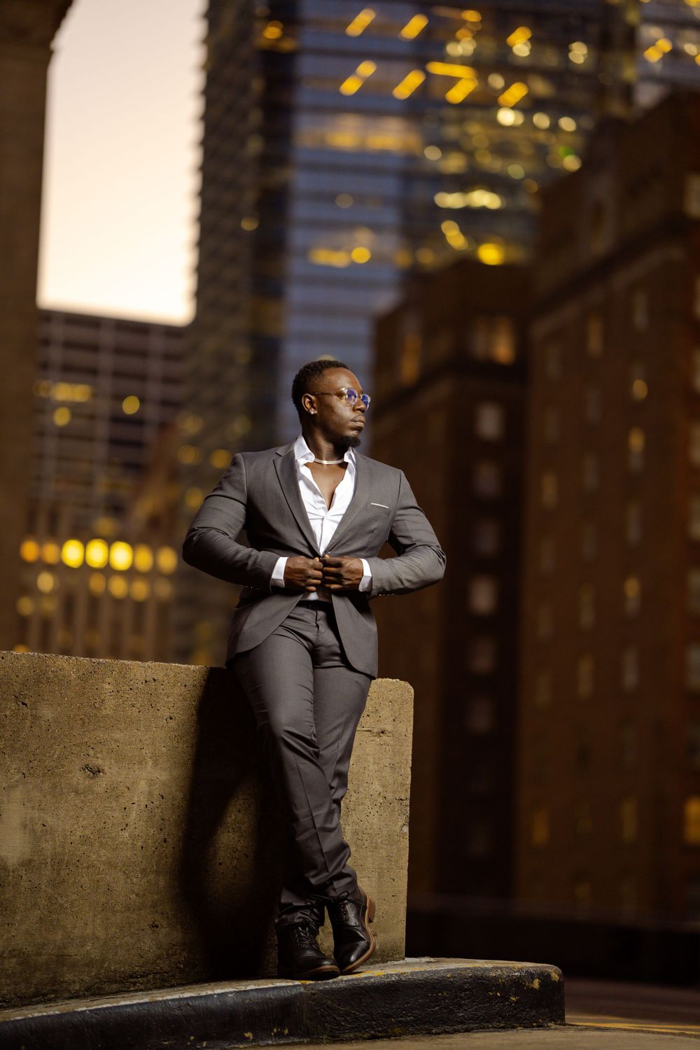 Professional in a gray suit poses confidently against an urban backdrop with glowing city lights at dusk.