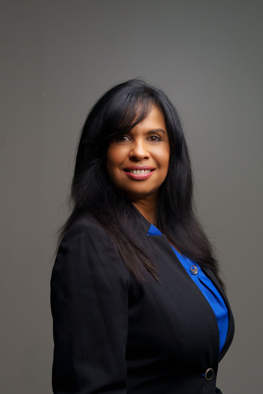 Professional headshot portrait of a person in a black blazer and blue blouse against a gray background.