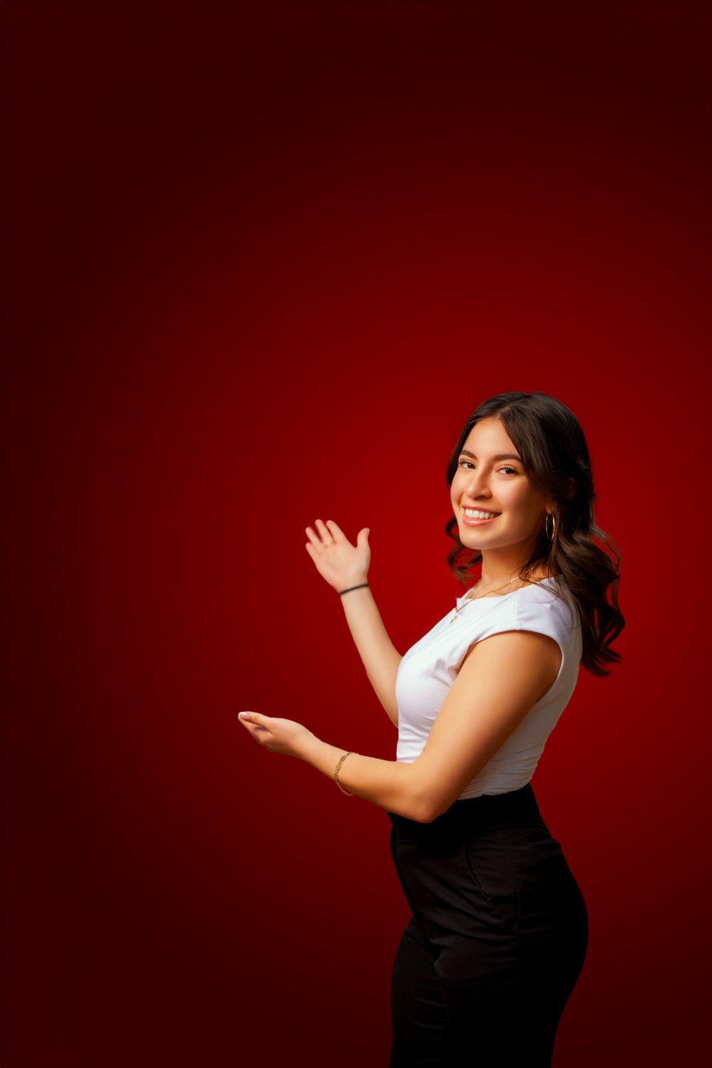 A person in a white t-shirt and black skirt poses with welcoming hand gestures against a deep red background.