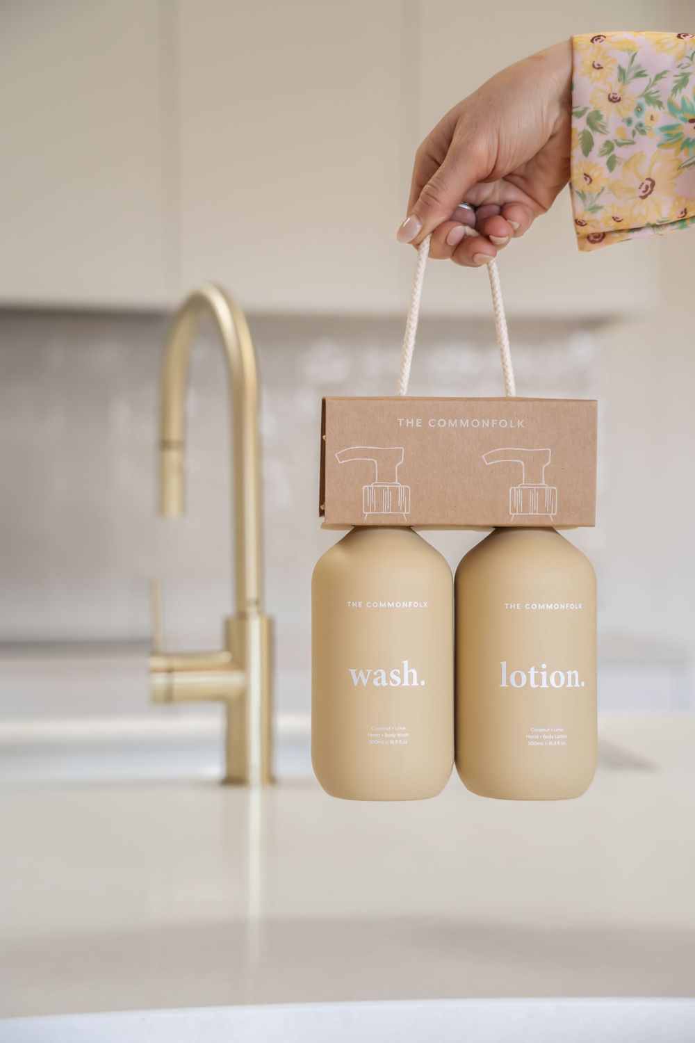 Hand holding beige pump bottles with brown packaging against white counter and gold faucet background.