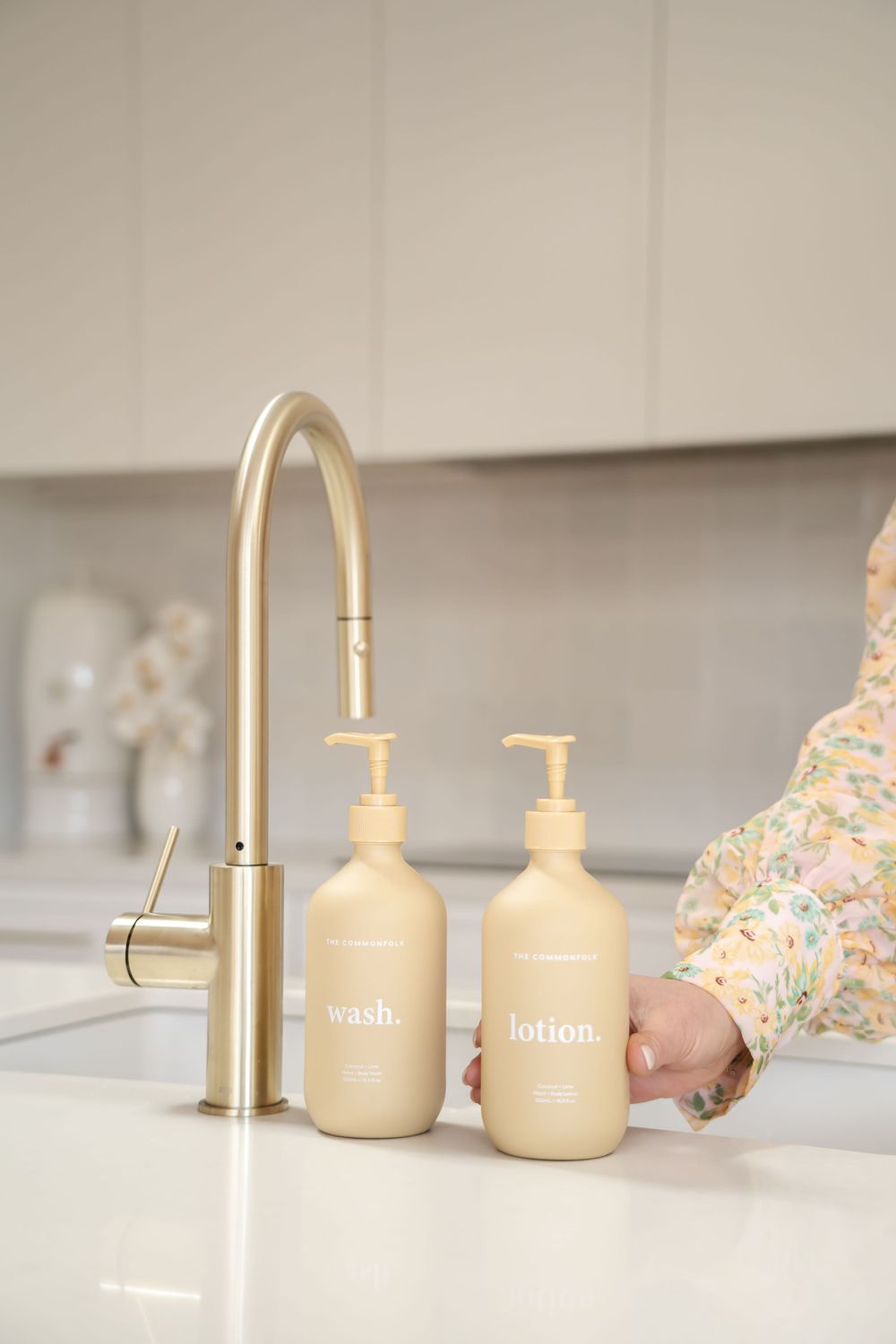 Minimalist bathroom setup featuring a gold faucet and beige pump bottles on white counter.