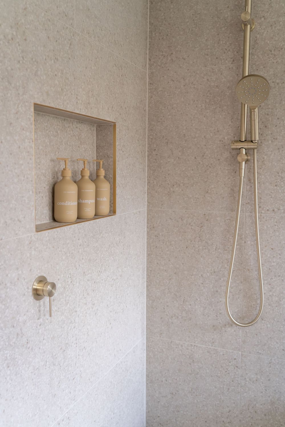 Modern shower interior with built-in niche storing beige pump bottles and gold fixtures.