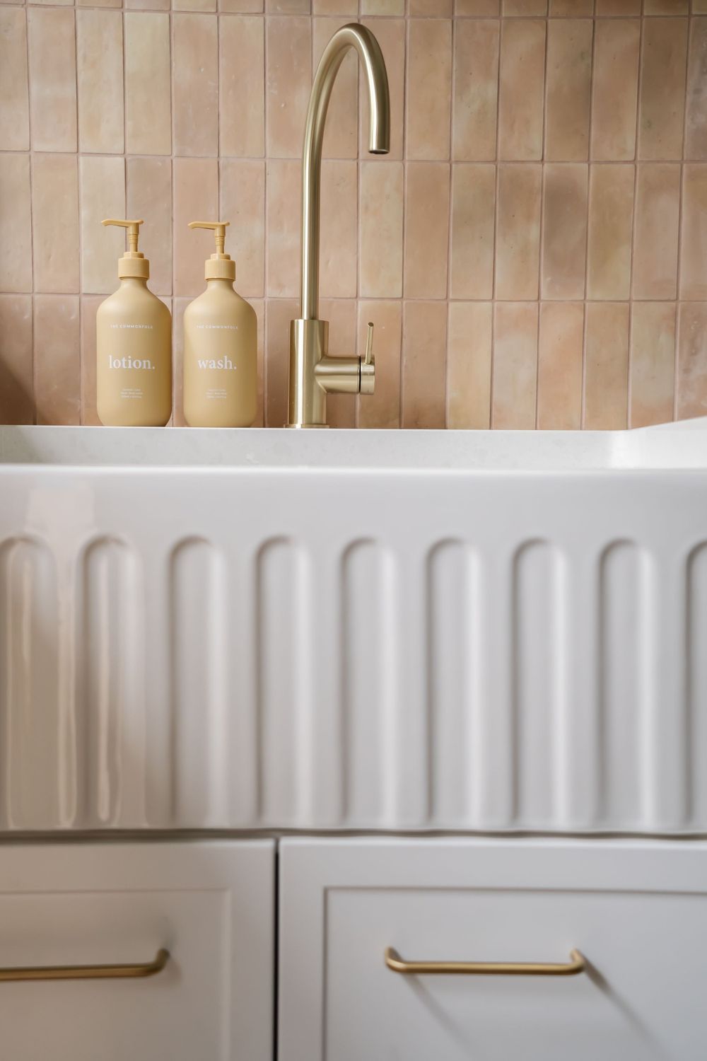 Modern bathroom sink with brass faucet and beige refillable dispensers against terracotta tile backsplash.