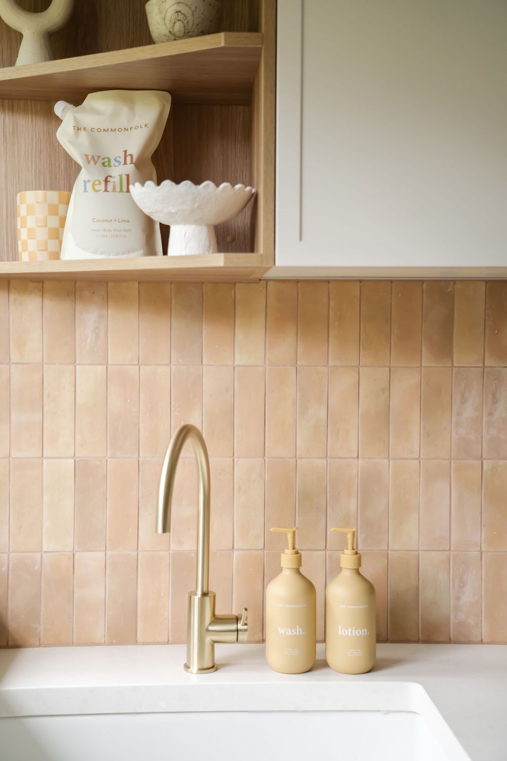Minimalist bathroom setup with brass faucet, refillable dispensers, and wooden shelving against peach tiles.