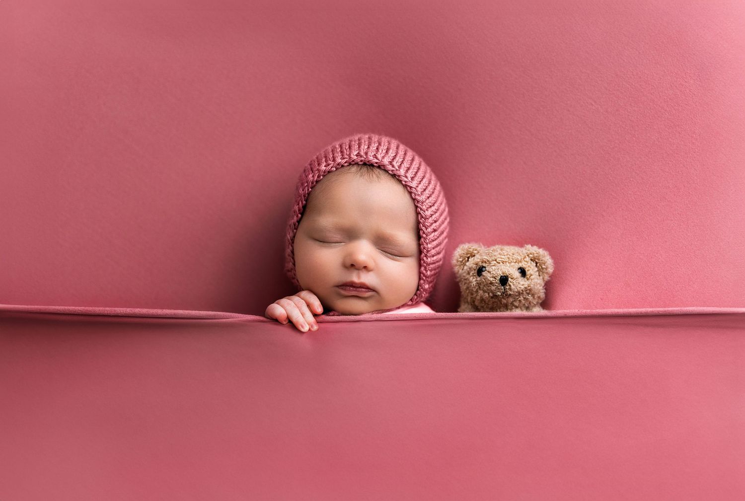 Sleeping baby wearing pink knit hat rests alongside small teddy bear against pink background.