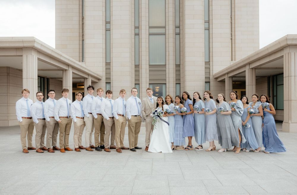 Large wedding party lineup with groomsmen in tan suits and bridesmaids in blue dresses outdoors.