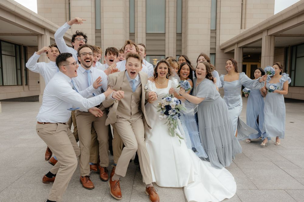 Excited wedding party surrounds laughing bride and groom in fun celebratory group photo outdoors.