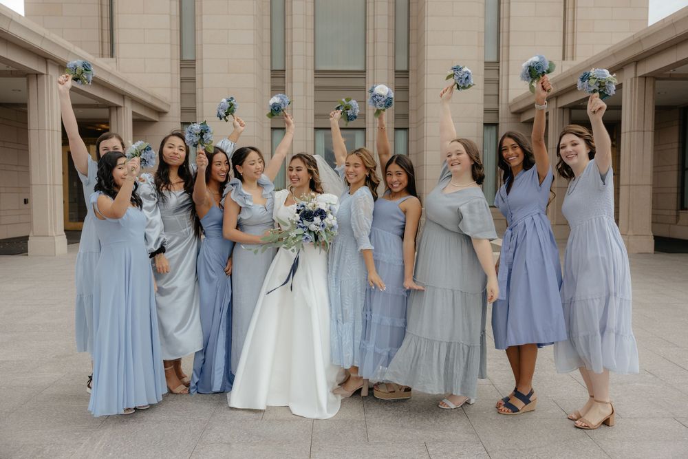 Bride surrounded by bridesmaids in mismatched blue dresses raising bouquets joyfully outdoors.