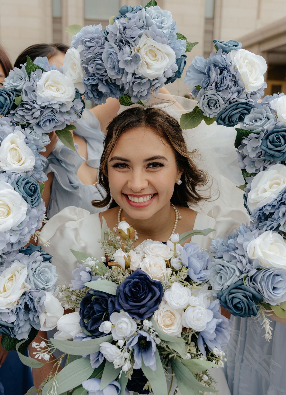 Smiling bride framed by blue and white floral bouquets held by bridesmaids in a circle.