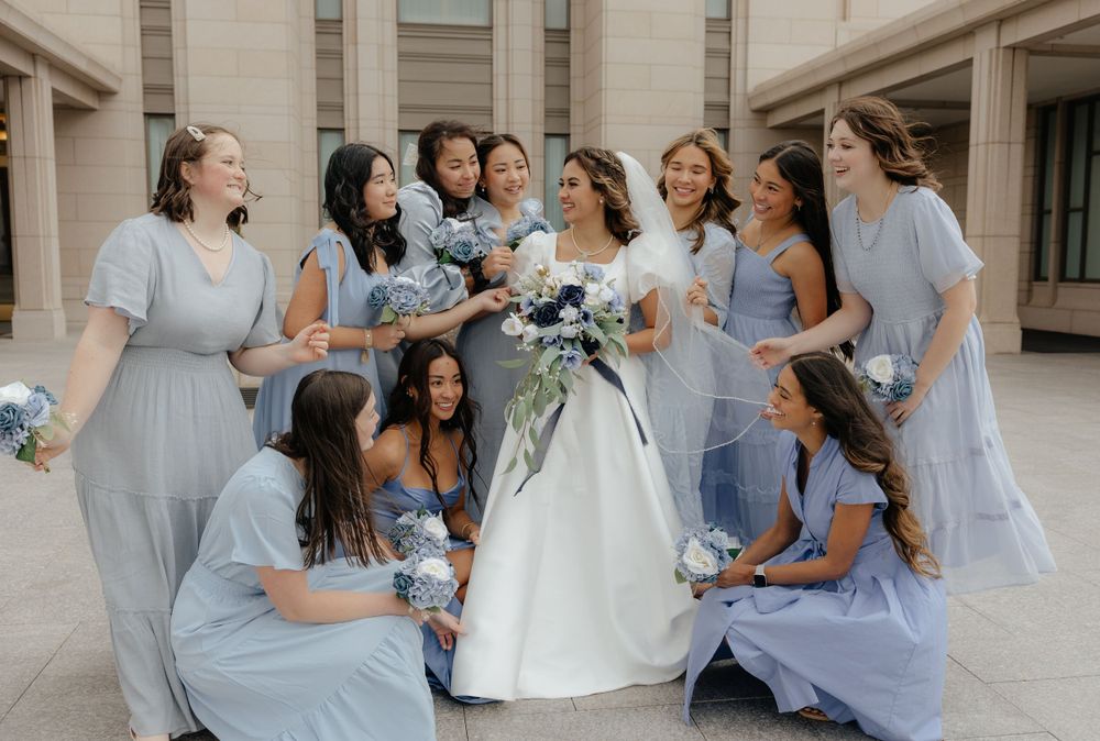 Bride in white gown laughing with bridesmaids in light blue dresses holding floral bouquets.