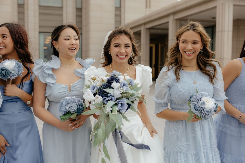 Bride smiling with bridesmaids in light blue dresses holding blue and white bouquets outside a temple.
