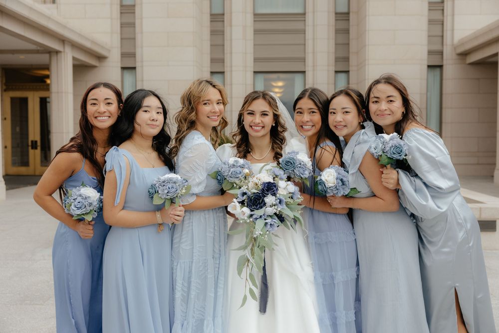 Bride and six bridesmaids in dusty blue gowns posing with floral bouquets outside a stone building.