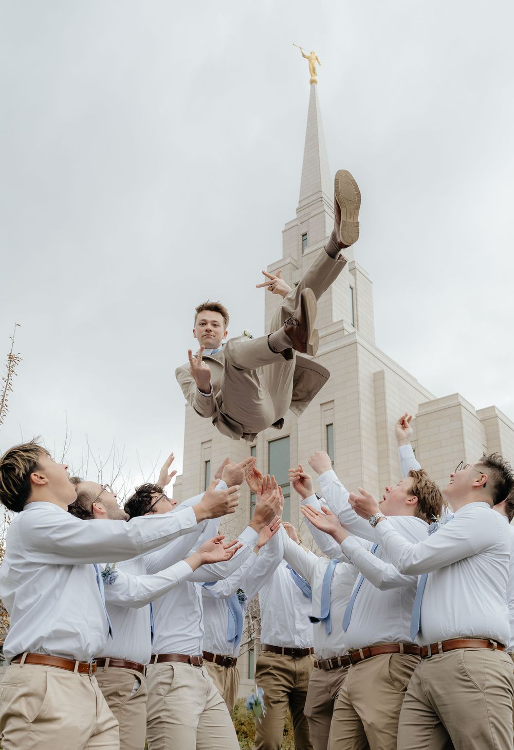 Groomsmen tossing groom in tan suit into the air in front of an LDS temple steeple.