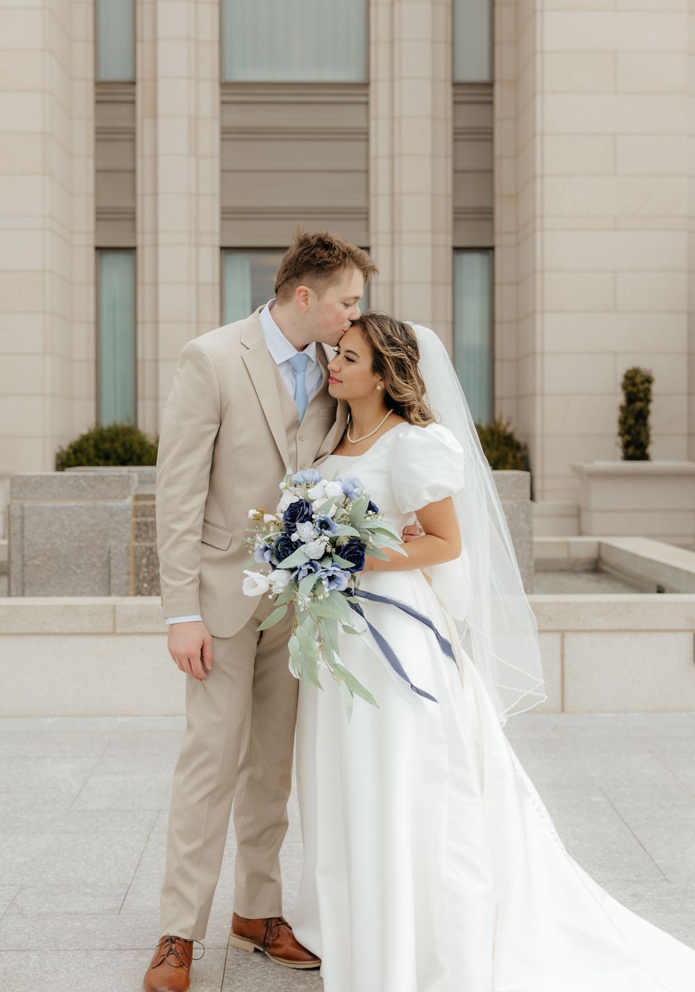 Bride and groom sharing a tender moment, she holds a blue and white bouquet outside a temple.