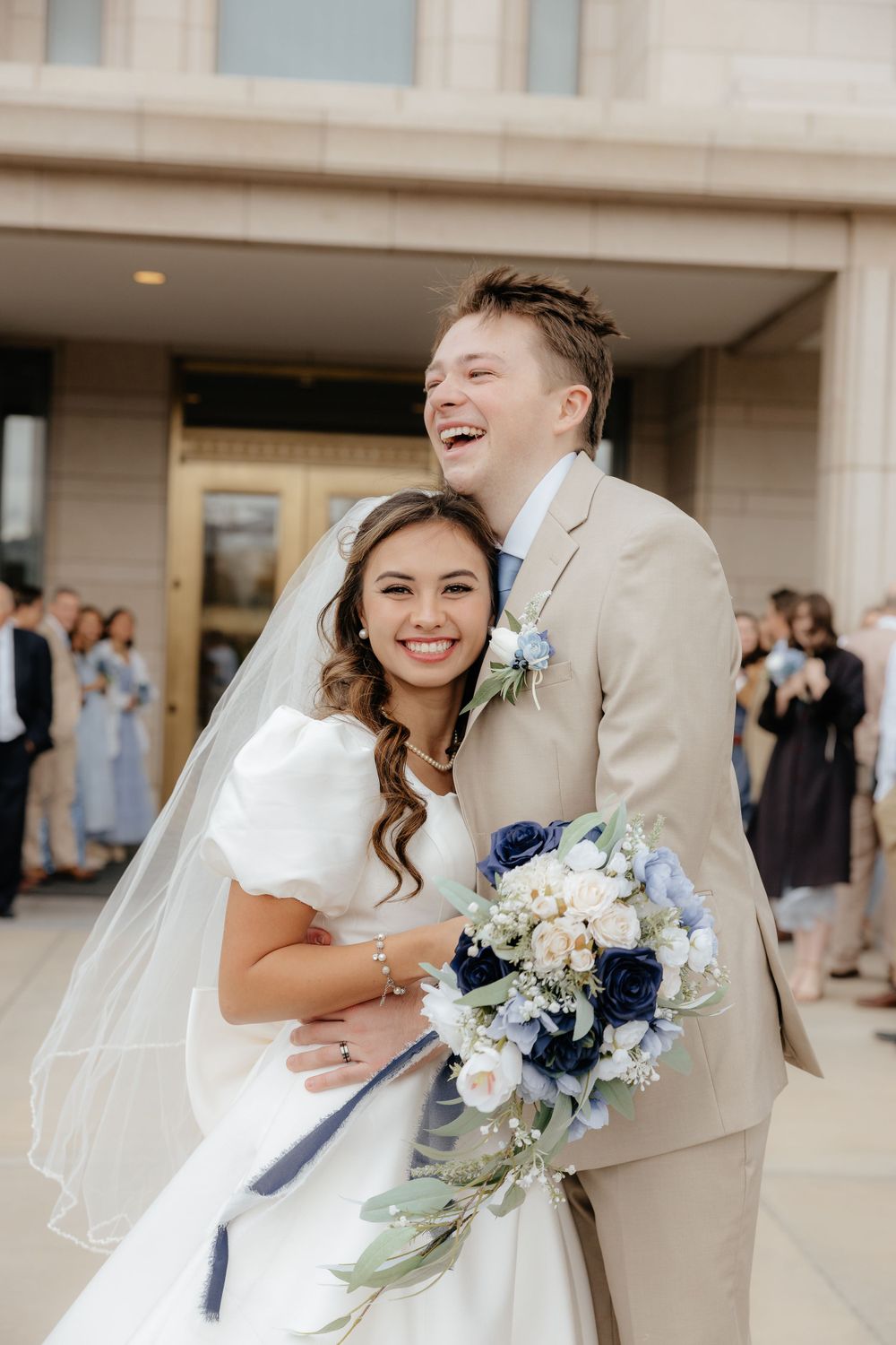 Happy bride and groom embrace outside LDS temple, bride holding blue and white bouquet.