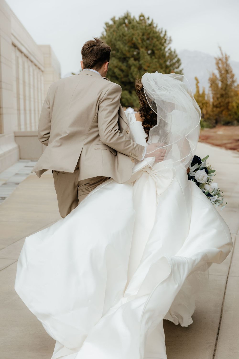 Bride and groom walking together outdoors, her white gown flowing in the wind near a elegant building.