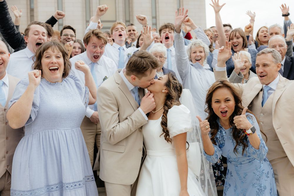 Joyful wedding party crowd cheering as bride and groom share a kiss on building steps.