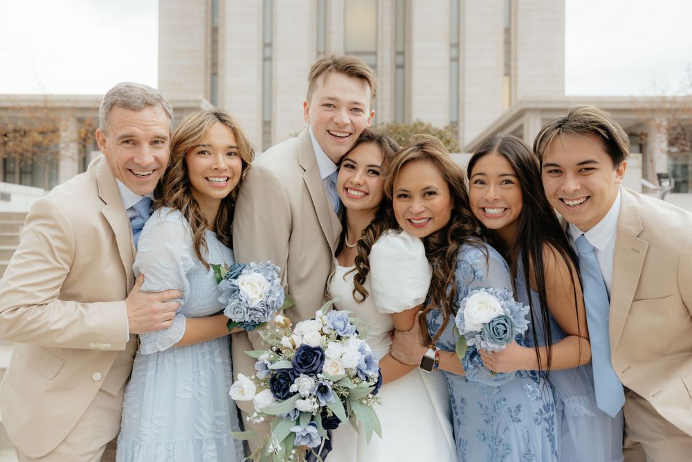 Bride and groom pose with wedding party in blue dresses and tan suits holding floral bouquets.