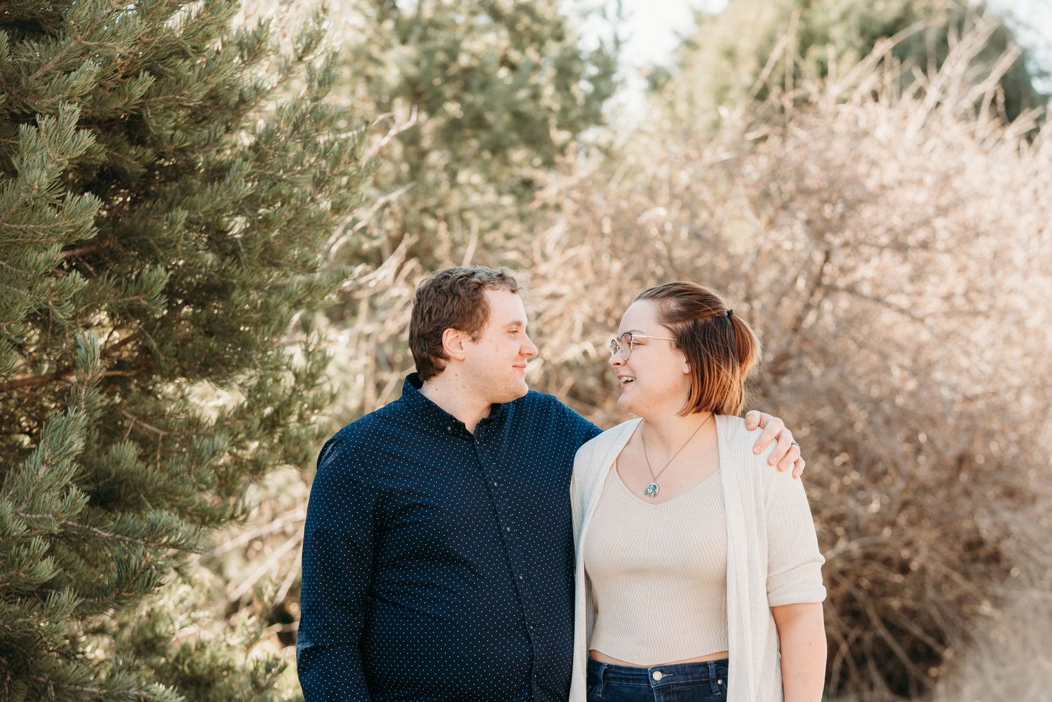 A couple shares a romantic gaze while standing outdoors against evergreen trees and winter foliage.