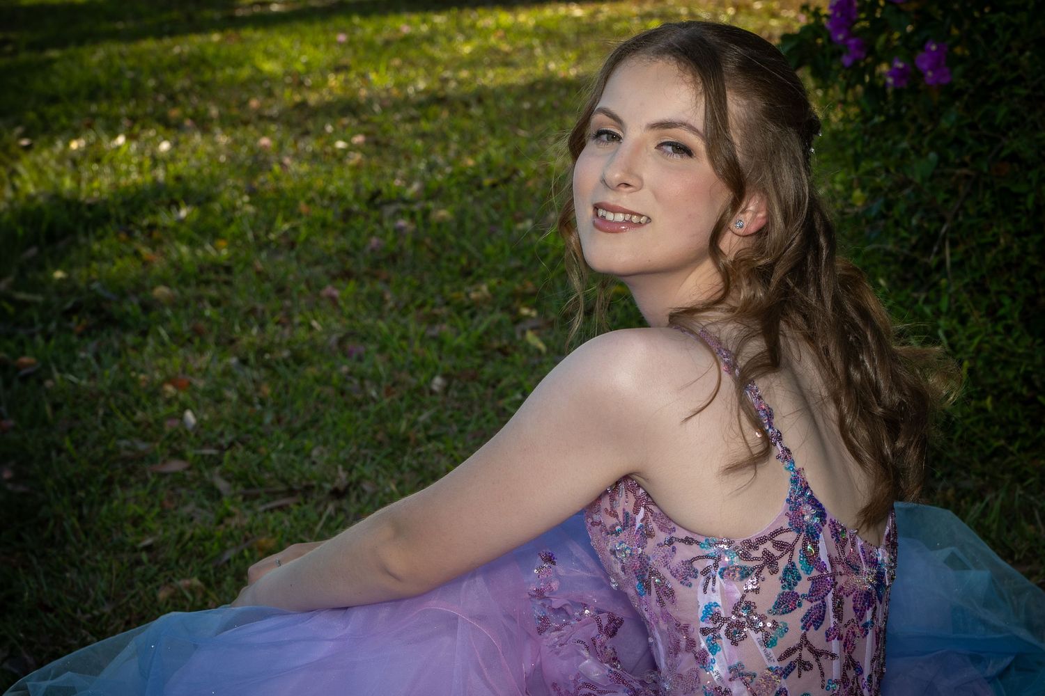 A woman in a flowing purple gown poses outdoors on grass with flowers in the background.