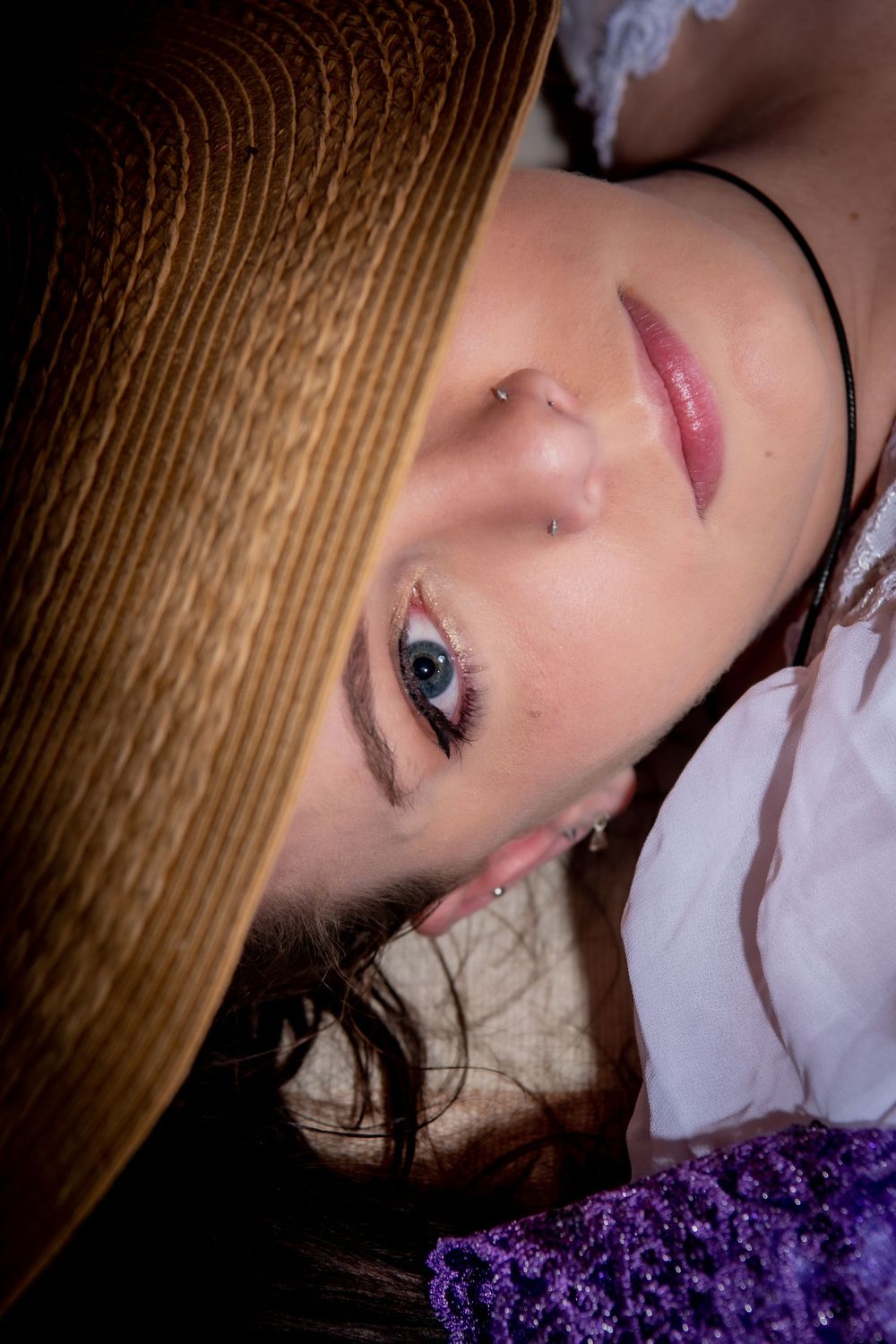 Close up beauty portrait with dramatic lighting wearing a straw hat against purple fabric background.