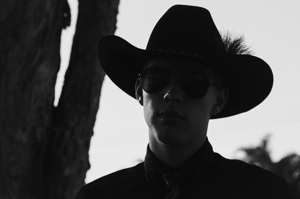 Black and white silhouette of a figure wearing a cowboy hat and sunglasses against a backlit background.