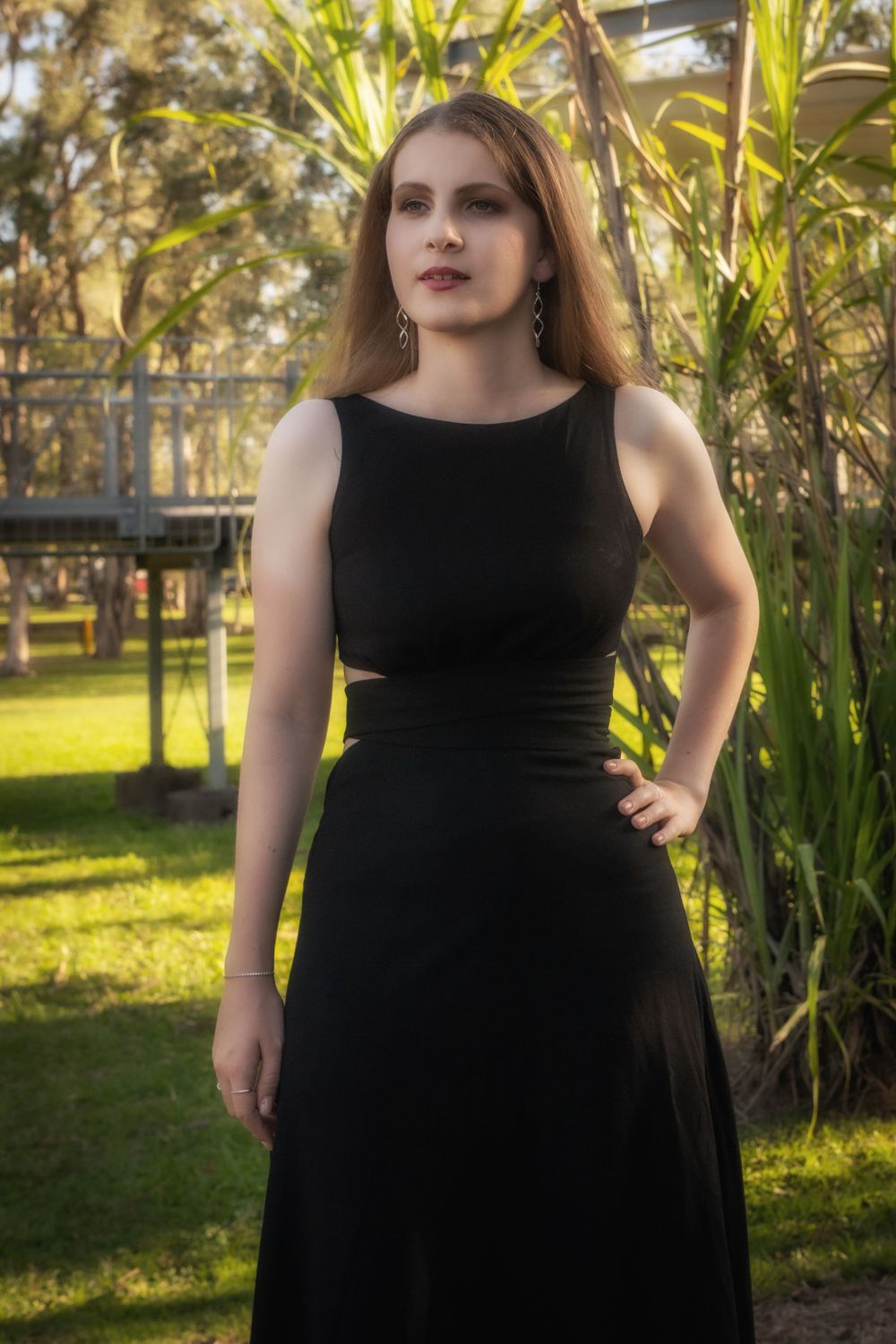A person stands in a garden wearing an elegant black sleeveless dress while golden sunlight filters through foliage.
