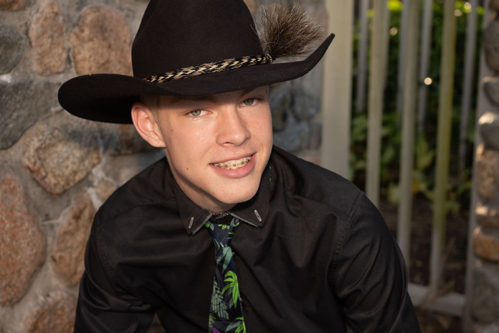 A person wearing a black western cowboy hat and black shirt with a patterned tie smiles against a stone wall background.