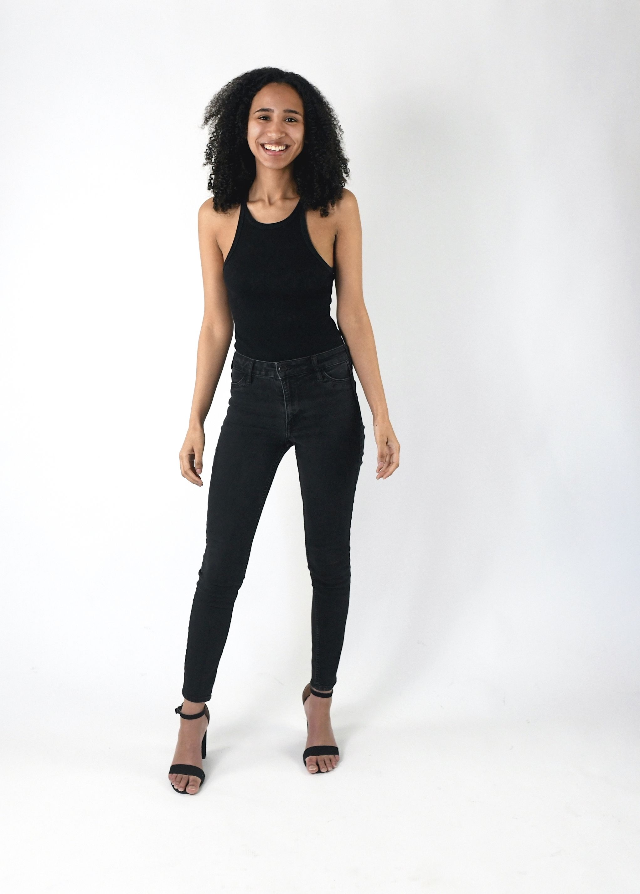 Woman in black tank top and jeans poses confidently against white background.